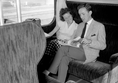 152859 Afbeelding van twee reizigers in een trein, met het tijdschrift Tussen de Rails van de N.S.